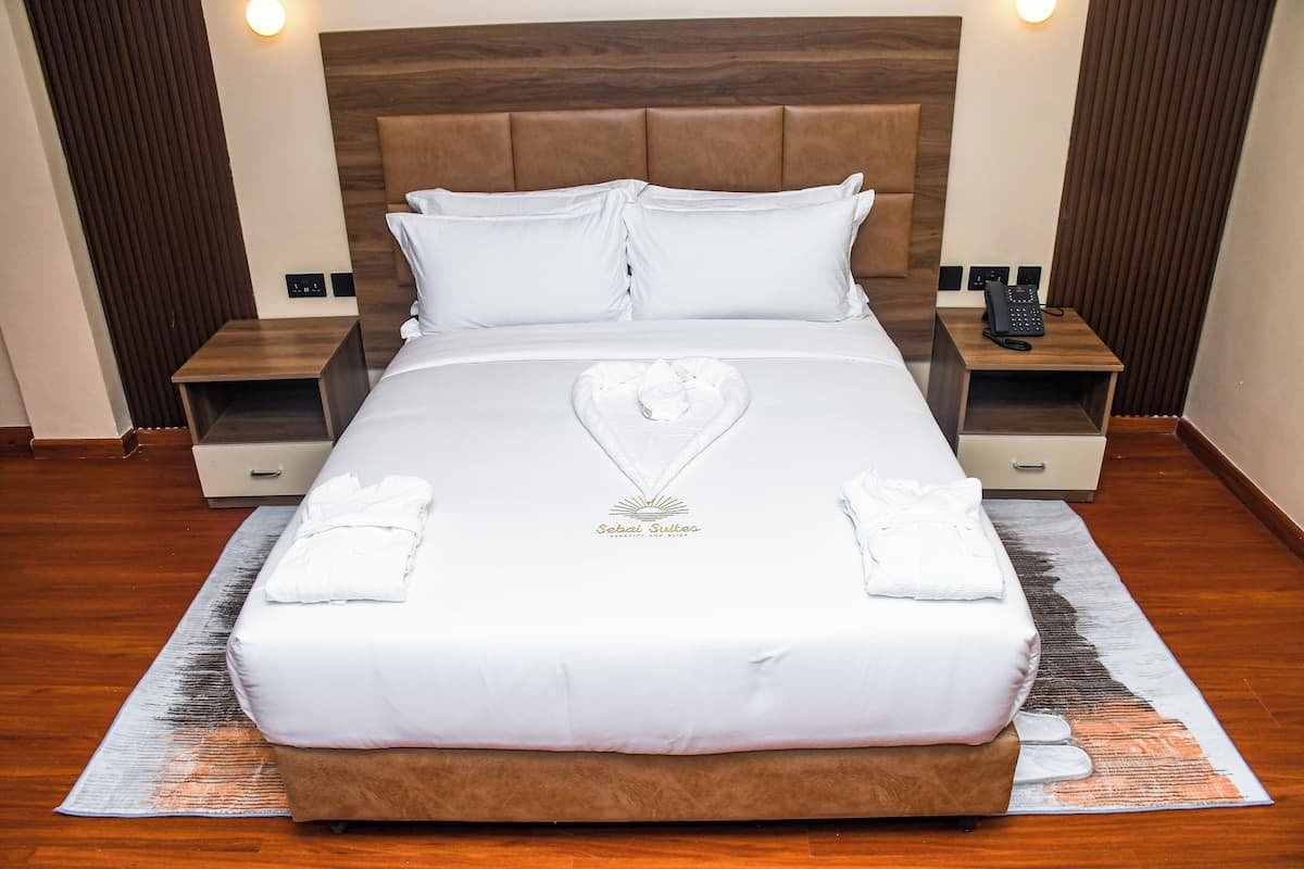 Cama bem arrumada com arranjo de toalha em formato de coração, cabeceira de madeira e mesas de cabeceira com abajures e telefone.