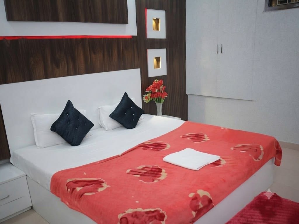 Un dormitorio con cama, almohadas, una colcha roja, un jarrón con flores y un armario.