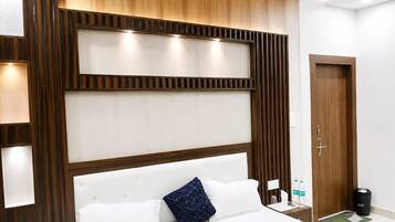 Habitación de hotel con cabecera de madera, cama con ropa blanca y almohada azul, mesita de noche con botellas de agua y puerta con panel de madera.