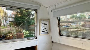 Een smalle hut met een raam dat uitkijkt over een gracht, een klein plankje met boeken en een lijst met bootafbeeldingen.