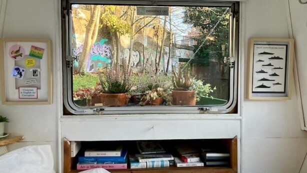 Een raam met uitzicht op bomen en een gebouw, een bed met wit beddengoed, een plank met boeken, en een lijst met een afbeelding aan de muur.