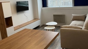 Moderni hotellihuone, jossa on puinen pöytä, sohva, seinälle asennettu televisio ja suuri ikkuna, josta on näkymä rakennuksiin.