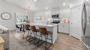 A modern kitchen with white cabinets, a central island, bar stools, a dining area with a table and chairs, and a large refrigerator.