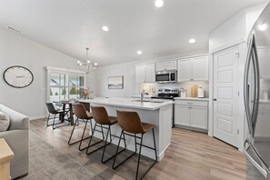 Una cocina moderna con armarios blancos, una isla central, taburetes de bar, un comedor con mesa y sillas, y un refrigerador grande.
