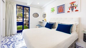 Un dormitorio con cama, una ventana con cortinas, una pared decorada con cuadros y una mesita con un florero.