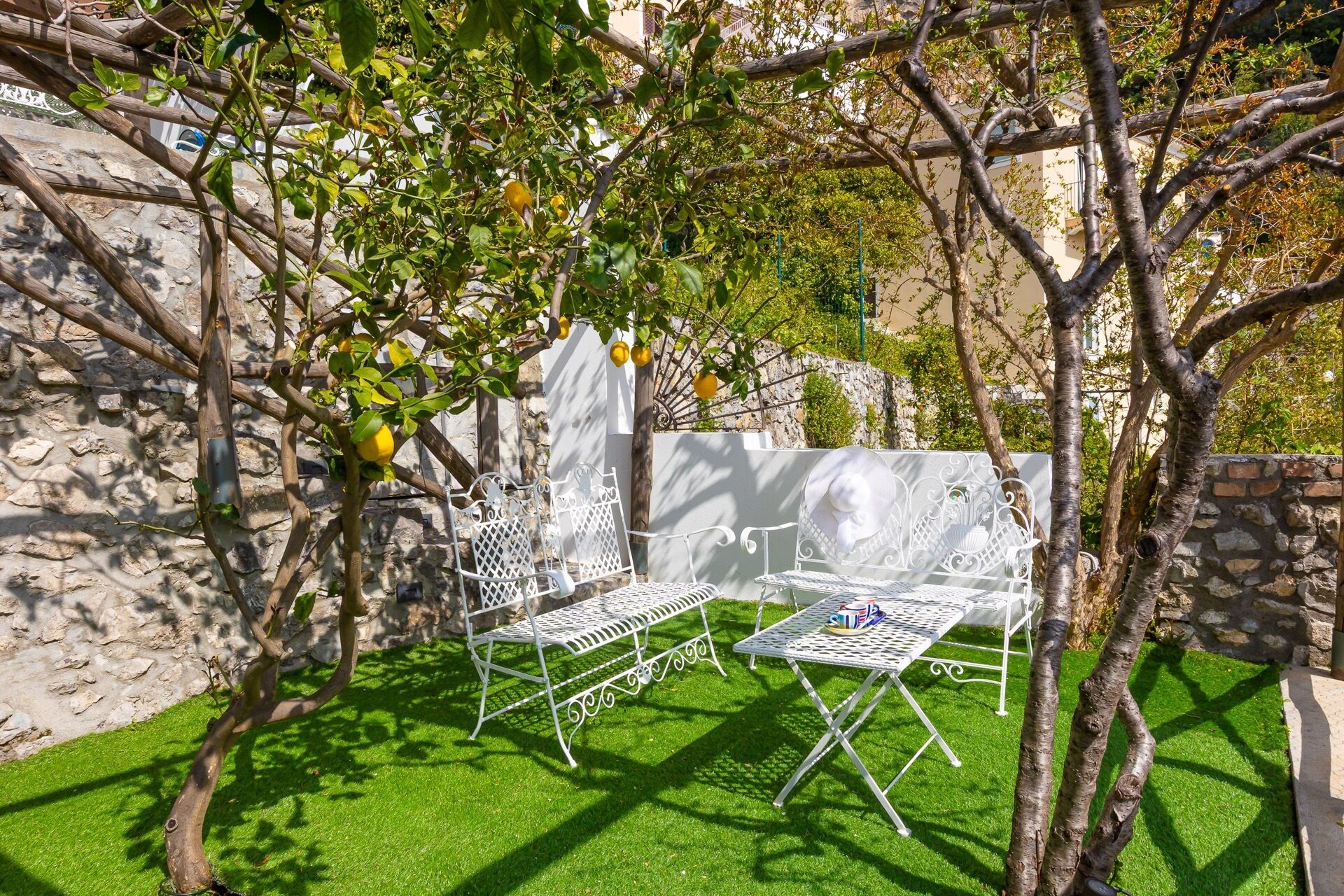 A garden with a white metal table and chairs, surrounded by trees and a stone wall.