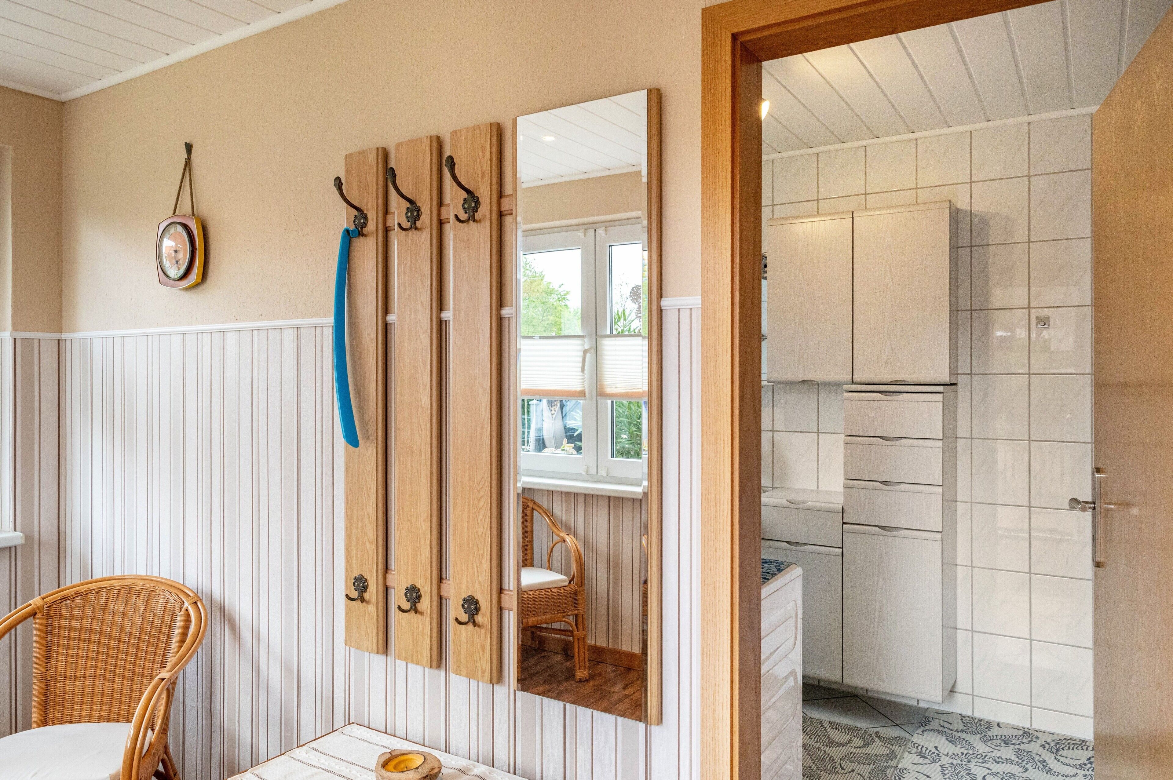 Une pièce avec un fauteuil en osier, un buffet en bois doté d’un miroir, et une vue sur une cuisine par une fenêtre.