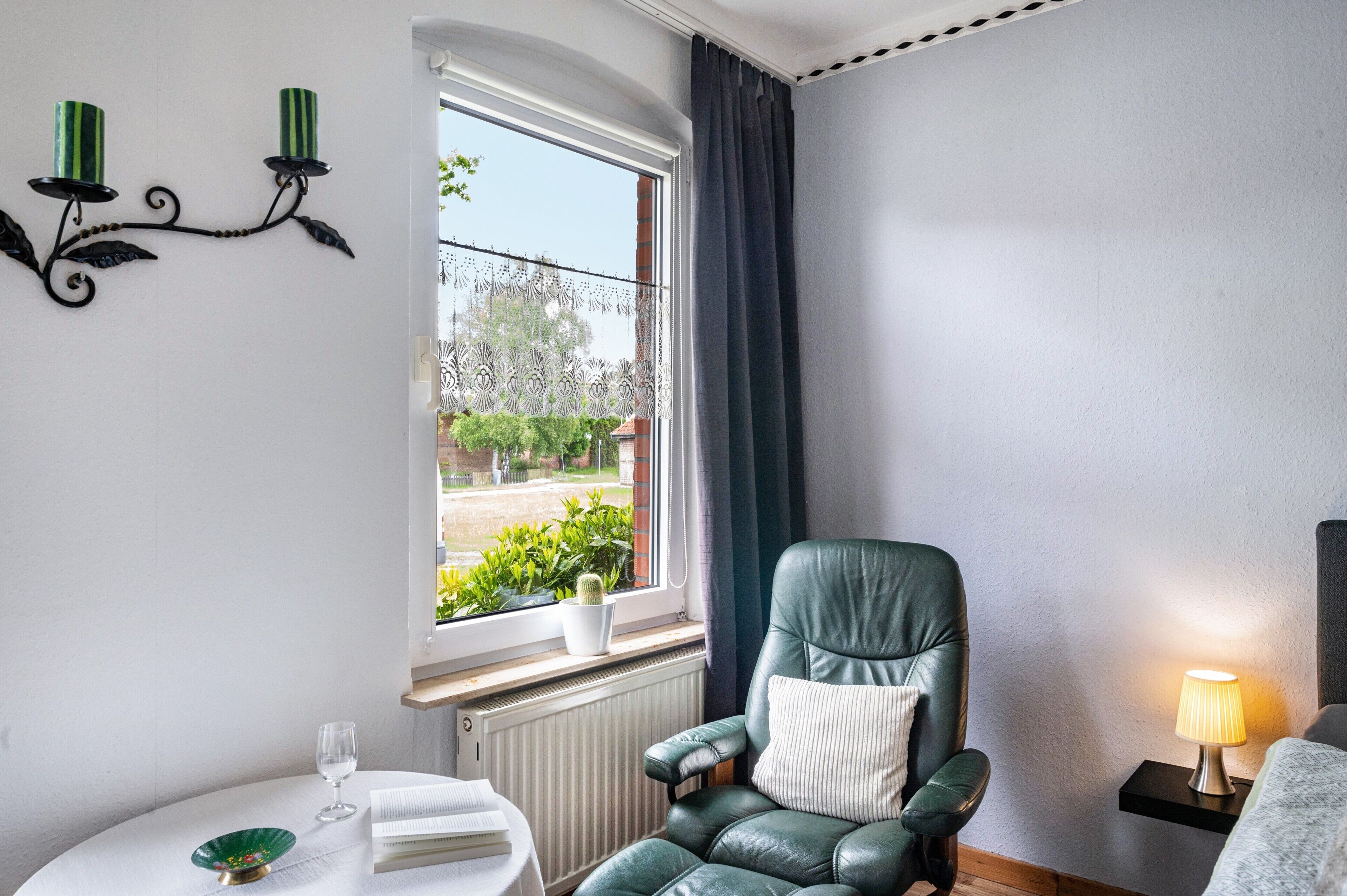 Ein Zimmer mit Fenster, ein grüner Ledersessel, ein Nachttisch mit Lampe und ein kleiner Topfpflanze.