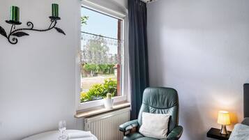 Ein Zimmer mit Fenster, ein grüner Ledersessel, ein Nachttisch mit Lampe und ein kleiner Topfpflanze.