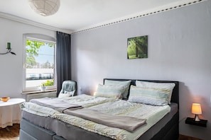 Une chambre à coucher avec un lit, une chaise, une table, une lampe, un tableau au mur et une fenêtre avec des rideaux.