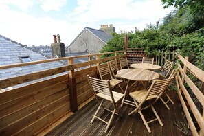 Eine Holzterrasse mit einem runden Tisch und Stühlen, umgeben von einem Holzzaun, mit Blick auf eine Wohngegend mit Häusern und einem klaren Himmel.