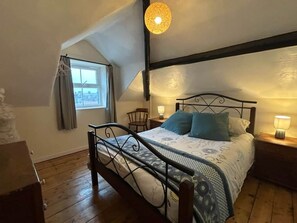 Ein Schlafzimmer mit schrägen Wänden, einem Bett mit Metallgestell, einem Nachttisch mit Lampe und einem Fenster mit Vorhängen.