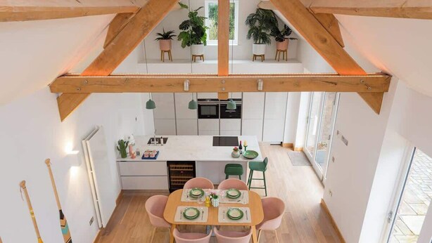 A dining area with a wooden table and chairs, a kitchen in the background, and potted plants on the upper level. - 2 Bed Converted Barn (Ombersley)