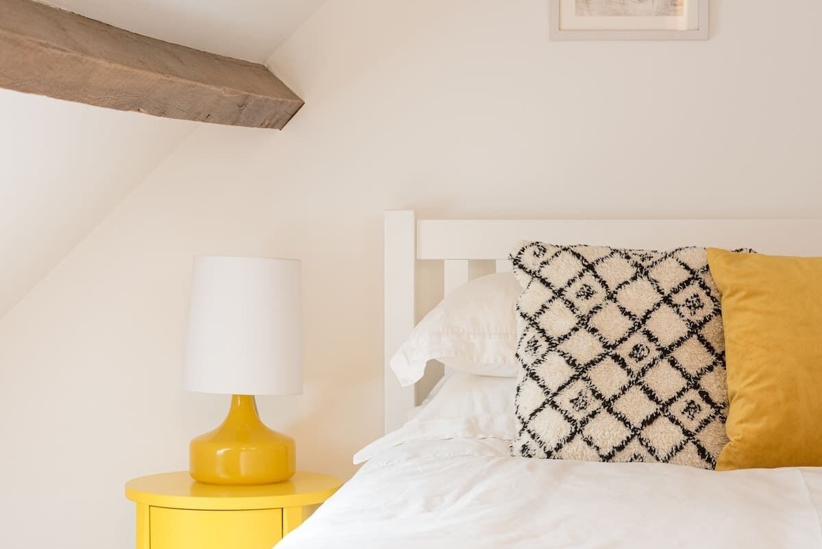 A bed with patterned pillows, a white headboard, and a yellow bedside table with a lamp.