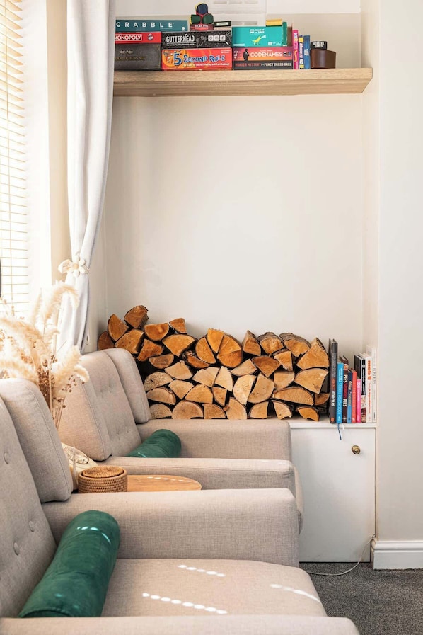 A living room with a sofa, a stack of firewood, a bookshelf with various books, and a window with curtains.