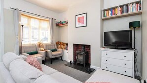 Een zithoek met een bank, fauteuil, open haard, televisie, boekenkast en een ingelijste foto aan de muur.