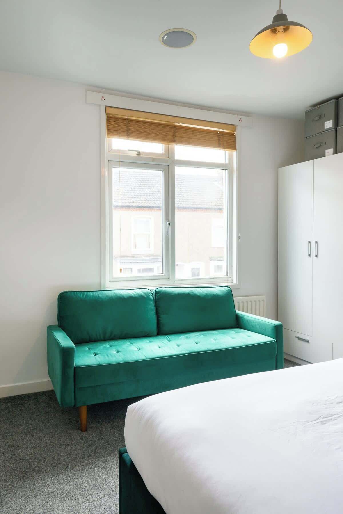 A room with a teal sofa, a white bed, a window with blinds, a radiator, and a ceiling light fixture.