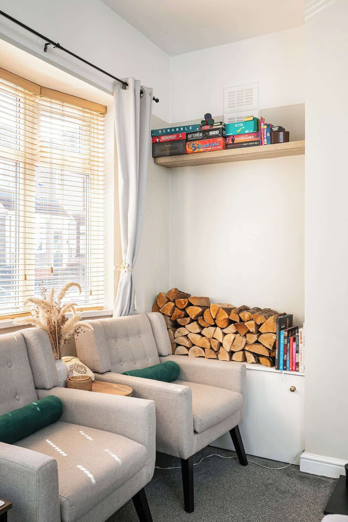 A living room with two armchairs, a window with blinds, a fireplace with a stack of logs, and a shelf with board games.