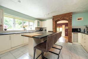 A modern kitchen with a central island, white cabinetry, and a brick archway leading to another room. - Spacious 5BR Home in Redditch (Redditch)