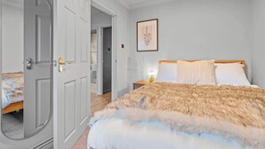 A bedroom with a large bed, a door, a framed picture on the wall, and a nightstand with a lamp. - Cosy 3 Bed with Parking (Rugby)