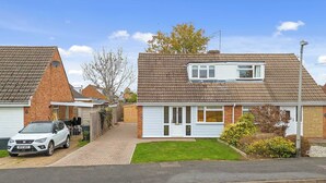 A residential house with a car parked in the driveway, a small lawn, and a clear sky. - Cosy 3 Bed with Parking (Rugby)