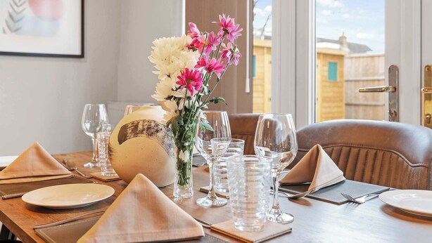 A dining table set with glasses, plates, and a vase of flowers. - Cosy 3 Bed with Parking (Rugby)
