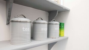 Tre contenitori grigi etichettati 'tè', 'zucchero' e 'caffè' su uno scaffale bianco.