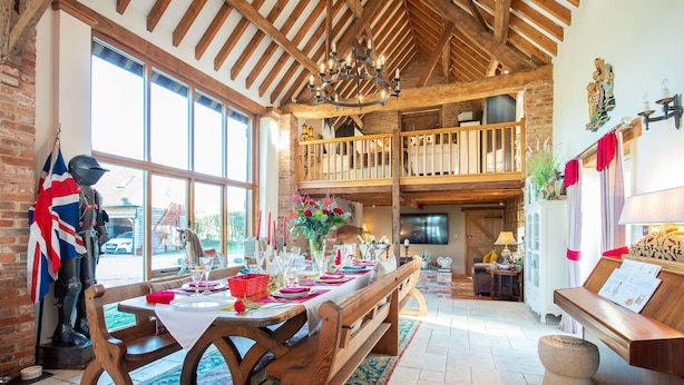 Un espace repas avec une longue table en bois dressée pour un repas, un balcon doté de balustrades en bois et un intérieur rustique avec des poutres apparentes et des murs en briques.