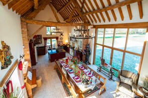 A spacious dining area with a long table, wooden beams on the ceiling, and a view of the water outside.