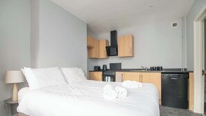 A hotel room with a bed, a bedside table with a lamp, a kitchenette with a stove, and a compact refrigerator. - F1 Stratford Studio Flat (Stratford-Upon-Avon)