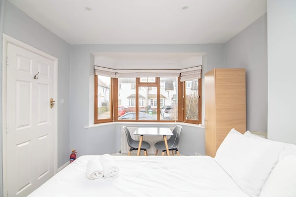 Une chambre à coucher avec un lit, un coin fenêtre avec deux fauteuils, une armoire blanche et une vue sur la rue.
