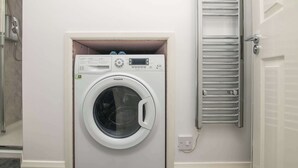 A compact, white wall-mounted washing machine with a control panel, a small shelf above it, and a metal towel rack to the right. - F1 Stratford Studio Flat (Stratford-Upon-Avon)