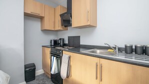 A compact kitchen with wooden cabinets, a black range hood, a stainless steel sink, and a black trash bin. - F1 Stratford Studio Flat (Stratford-Upon-Avon)