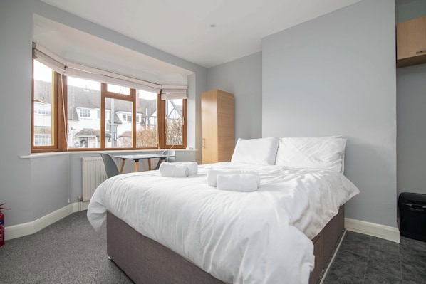 A bedroom with a bed, a desk, and a window with a view of houses.