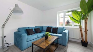 A living room with a blue sofa, a wooden coffee table, a floor lamp, and a large plant. - Compton House - 4 Bed (Cheltenham)