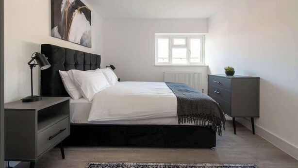 A bedroom with a bed, bedside tables, a radiator, a window, and a painting on the wall. - Compton House - 4 Bed (Cheltenham)