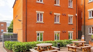 A brick building with multiple windows, outdoor wooden picnic tables, and a small garden area. - For Students Only - Park Court in Lincoln - Campus Accommodation (Lincoln)