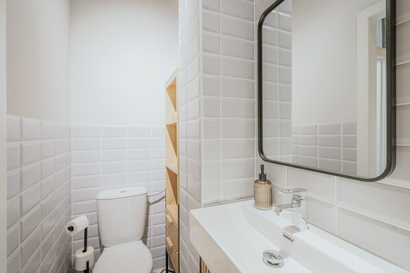 Un baño con paredes de azulejos blancos, un inodoro blanco, un lavabo con espejo y un dispensador de jabón.