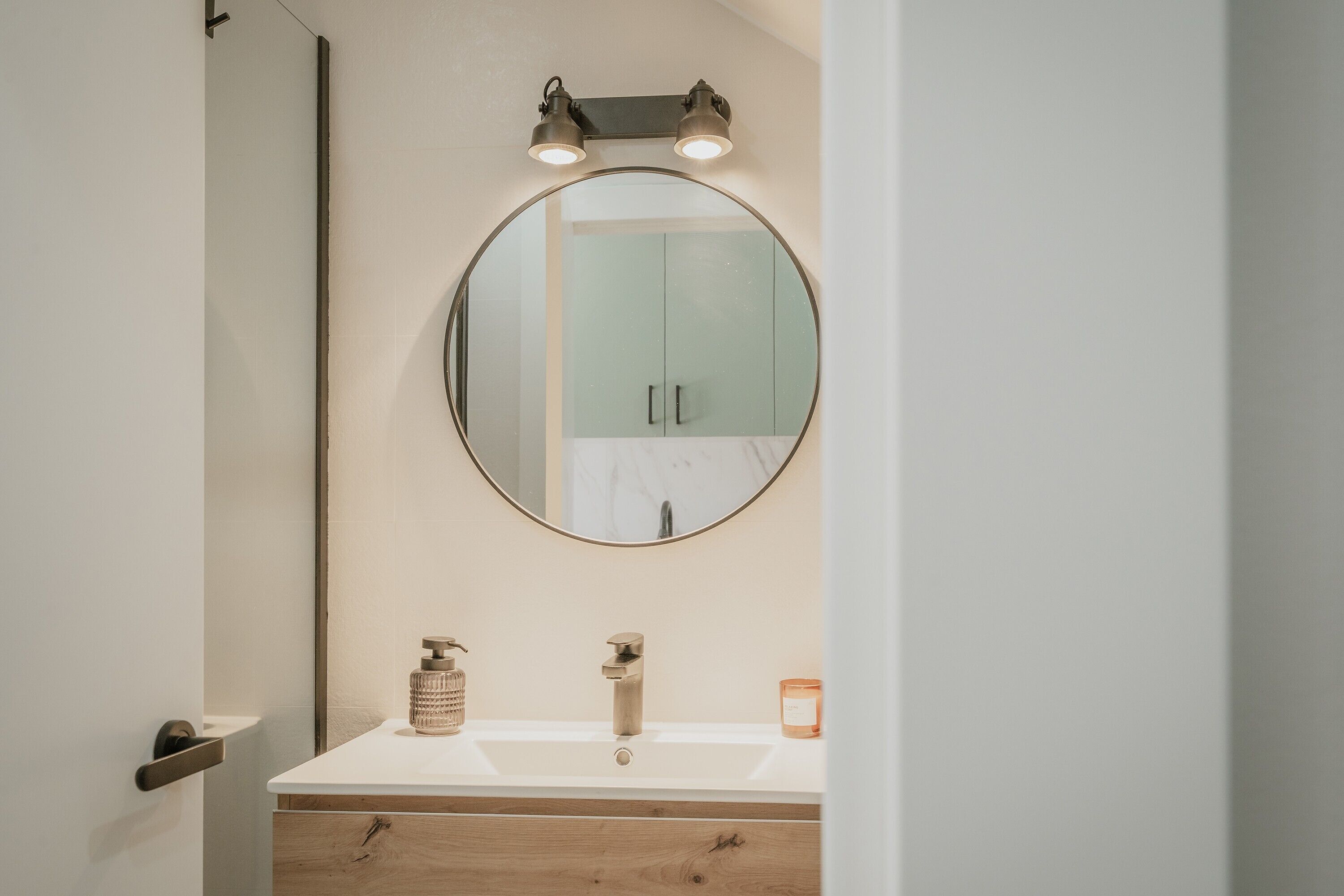 Un baño con un espejo redondo, un lavabo blanco, un mueble de tocador de madera y una puerta.