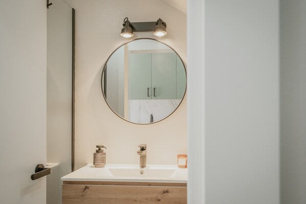 A bathroom with a round mirror, a white sink, a wooden vanity, and a door.