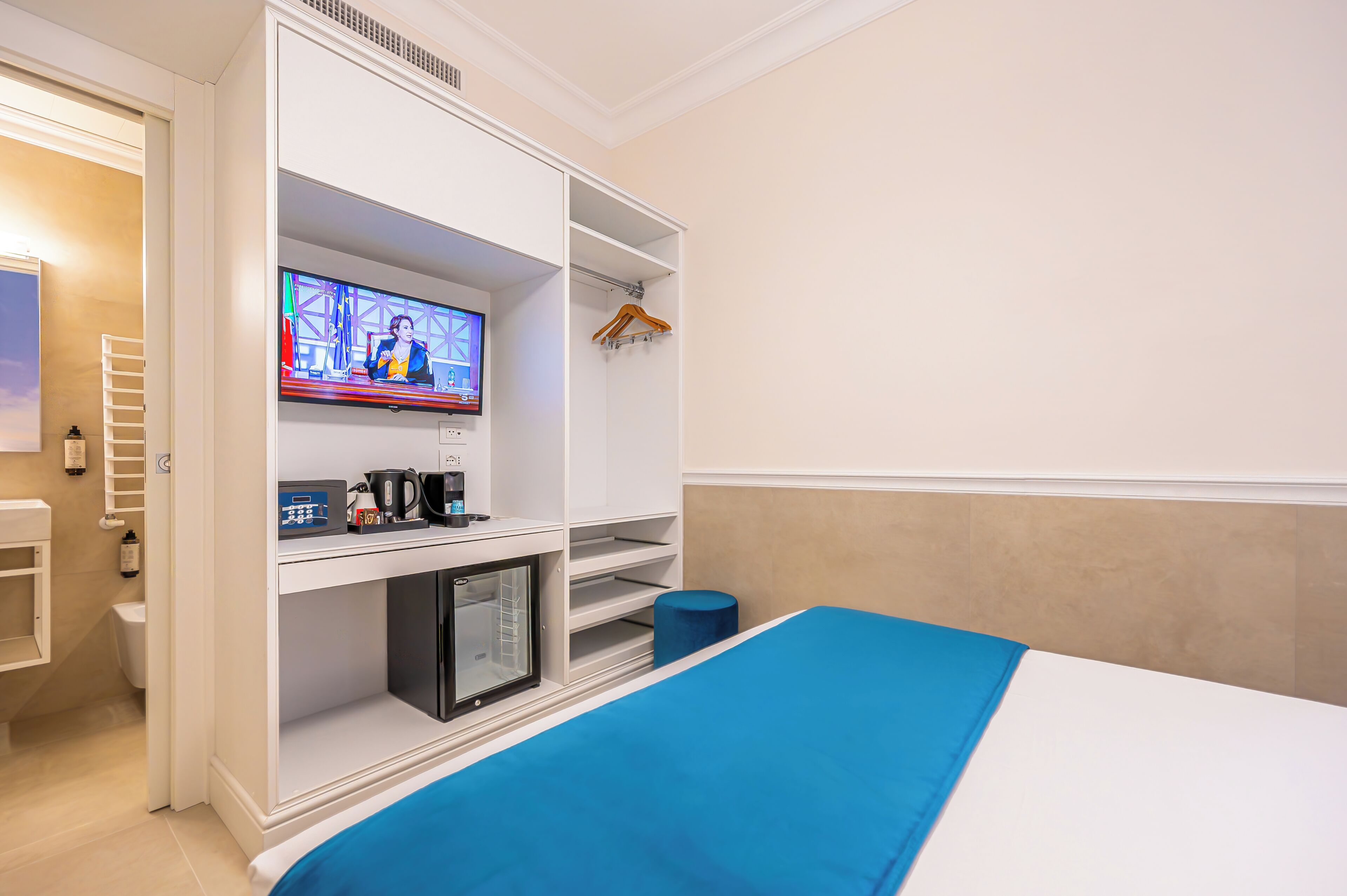 Une chambre d’hôtel avec un lit, un téléviseur fixé à une armoire blanche, une petite table avec une bouilloire et une machine à café, et une couverture de lit bleue.