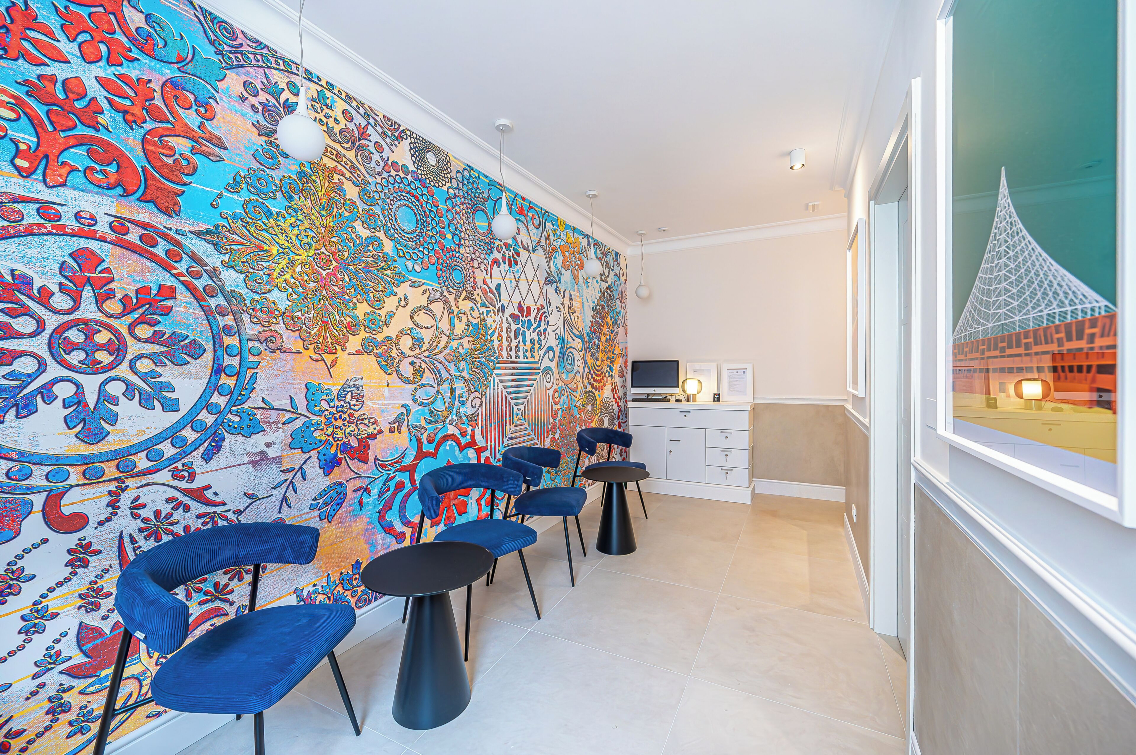 A room with a colorful tapestry, blue chairs, a small table, and a window with a view of a building.