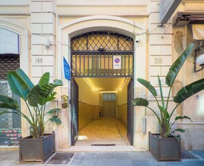 Entrada de um edifício com portão de metal, plantas em vasos e placa indicando área de estacionamento.