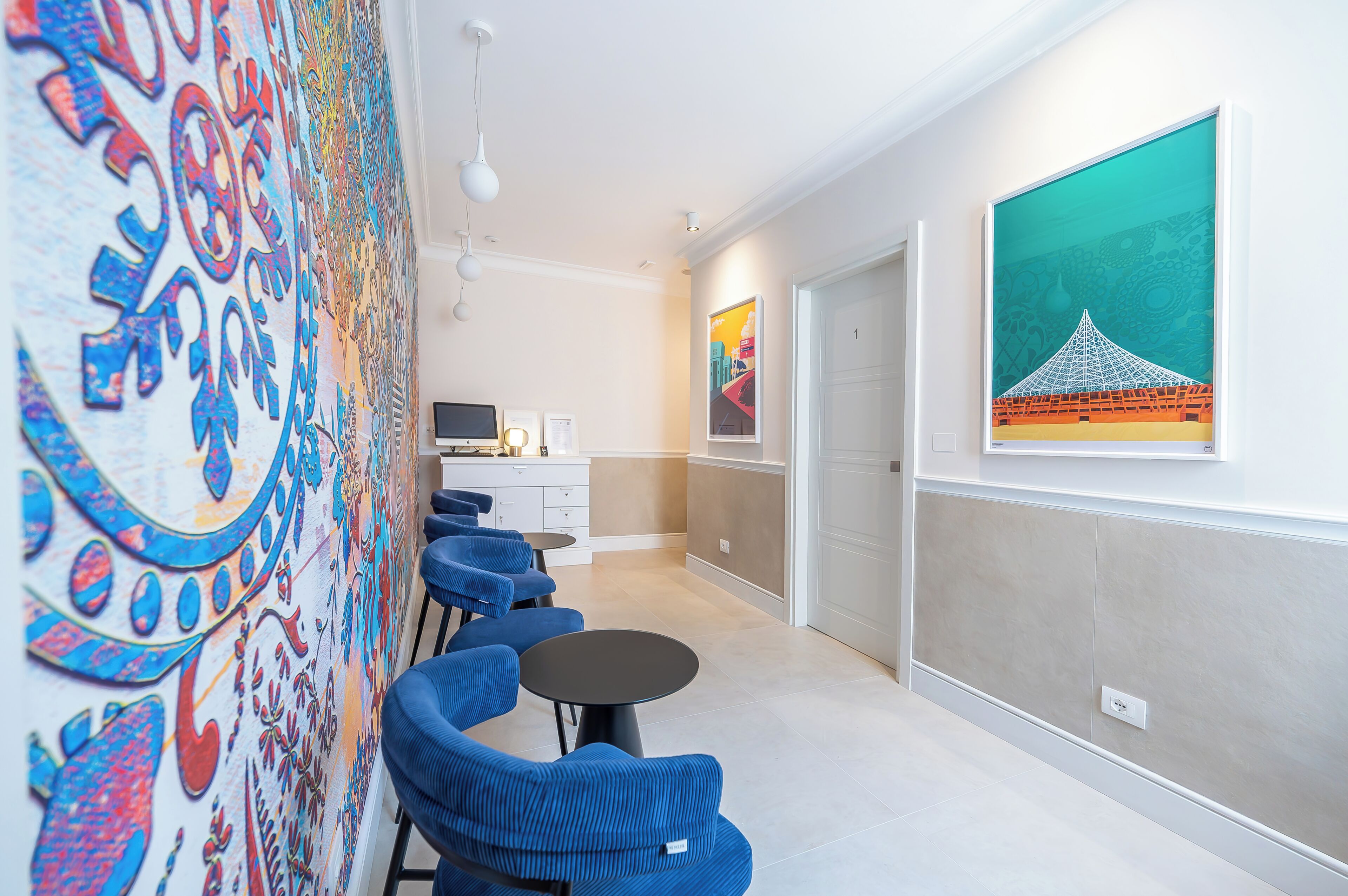 A hotel corridor with blue chairs, a dining table, a kitchen area, and artwork on the walls.