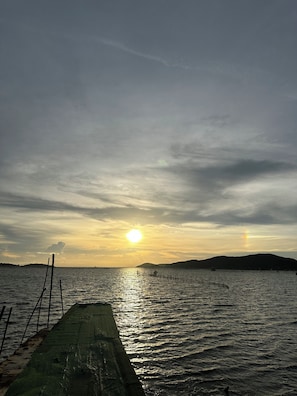 Vista para a frente do mar com um cais que se estende em águas calmas, um pôr do sol ao fundo e uma colina distante.