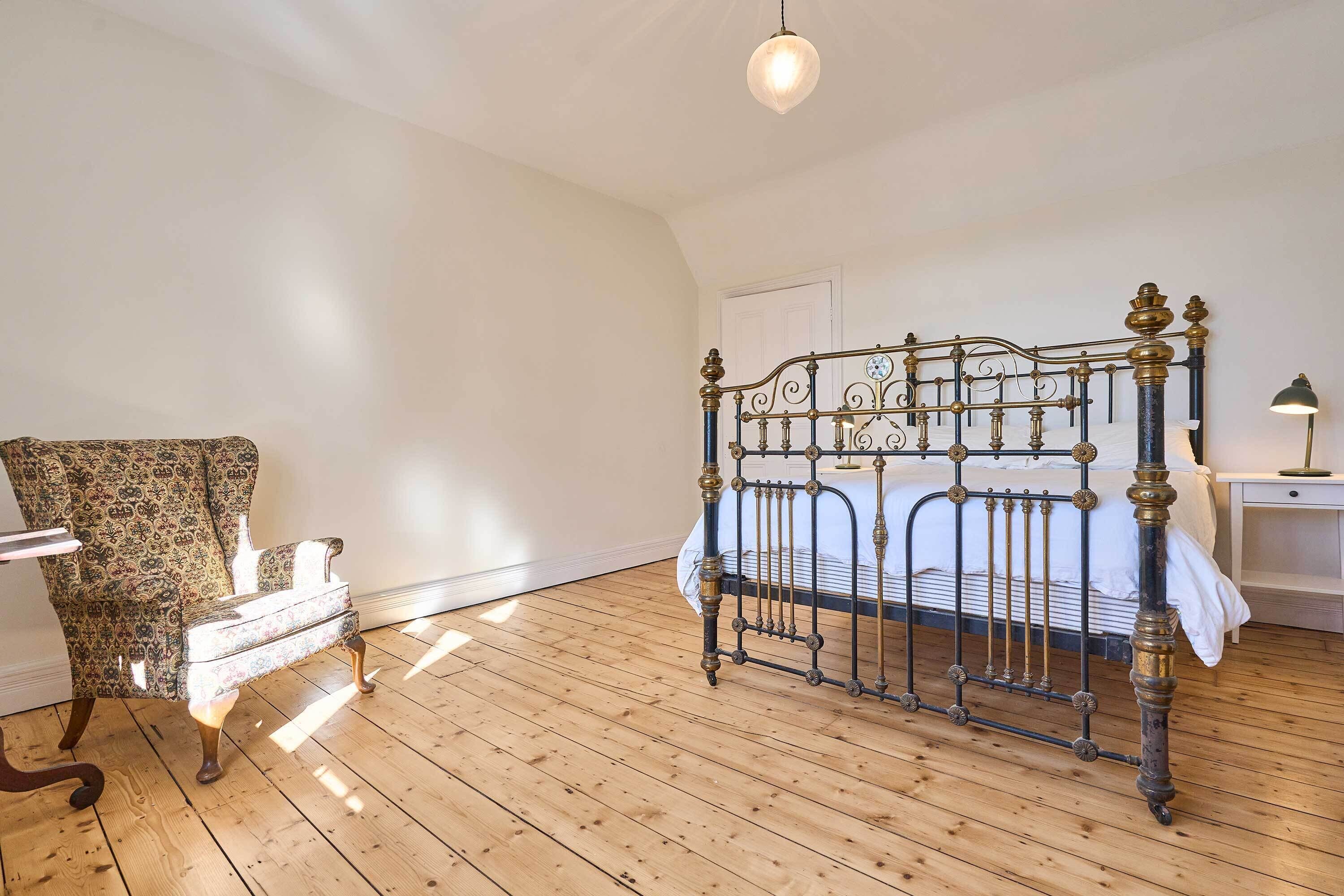 A room with a wooden floor, a bed with a metal frame, a patterned chair, and a bedside table with a lamp.
