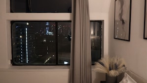 Ein Zimmer mit großem Fenster und Blick auf eine nächtliche Stadtlandschaft, einem Vorhang, einem gerahmten abstrakten Kunstwerk und einer Zimmerpflanze.