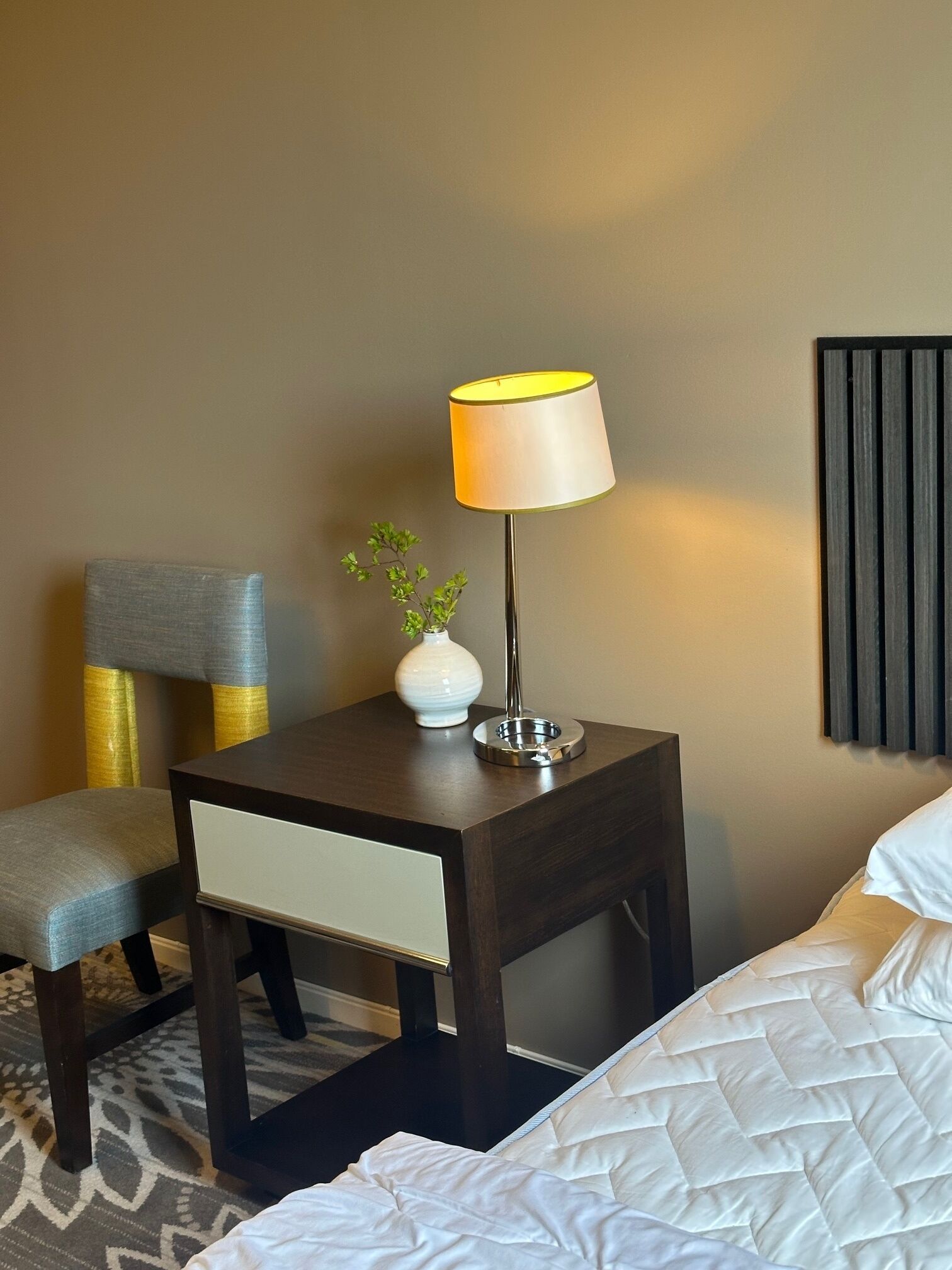Une chambre d’hôtel avec un lit, une table de chevet avec une lampe, un vase avec une plante et une chaise.