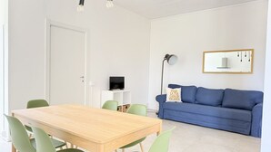 A modern living room with a wooden dining table, green chairs, a blue sofa, a TV, and a wall-mounted mirror. - PP11 - Potenza Picena, new three-room apartment with pool and air conditioning (Potenza Picena)