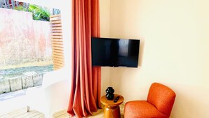 A modern living room with a flat-screen TV, a red armchair, a wooden side table, and a large window with curtains. - Studio Colibri - Terrace 2 pers, 3mn from Anse Mitan beach (Les Trois-Îlets)